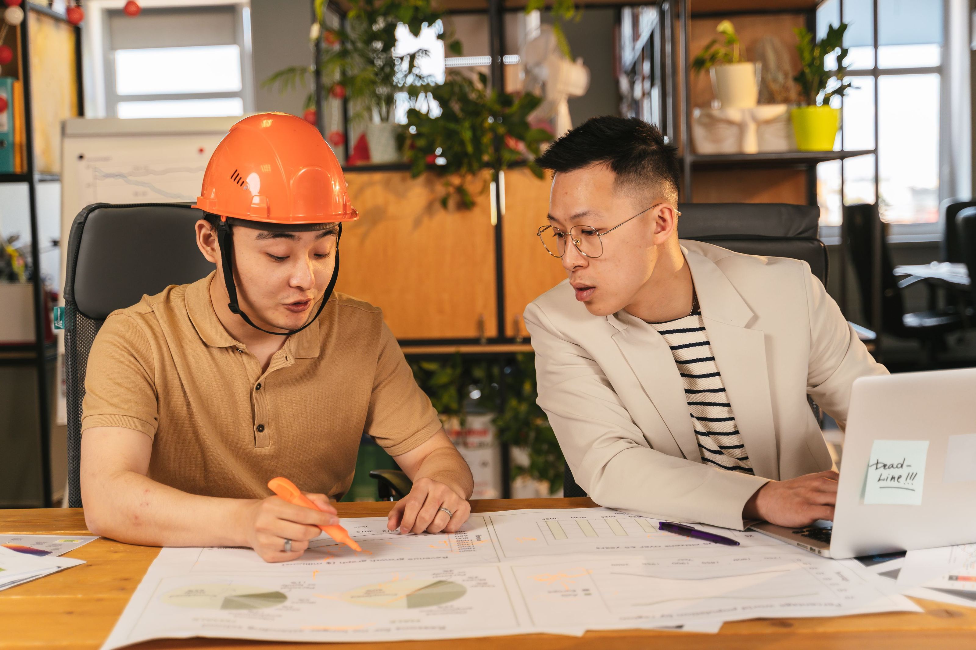 Two colleagues, one in a hard hat, reviewing plans and a laptop at an office desk