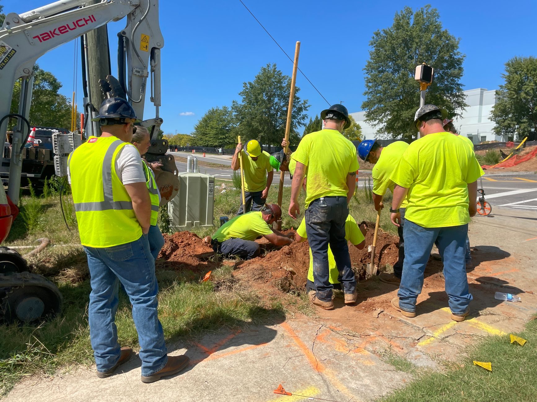 A utility crew in high-visibility vests standing at an excavation beside a compact excavator